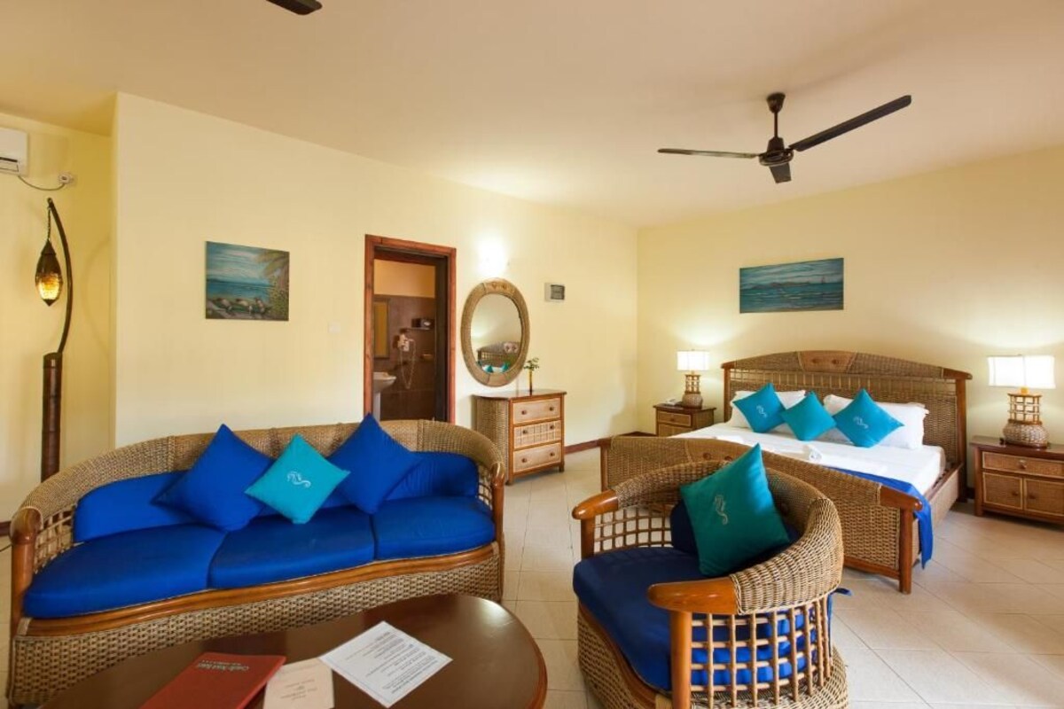 A room with a bed, two wicker sofas with blue cushions, a wooden table, a ceiling fan, and a mirror on the wall.