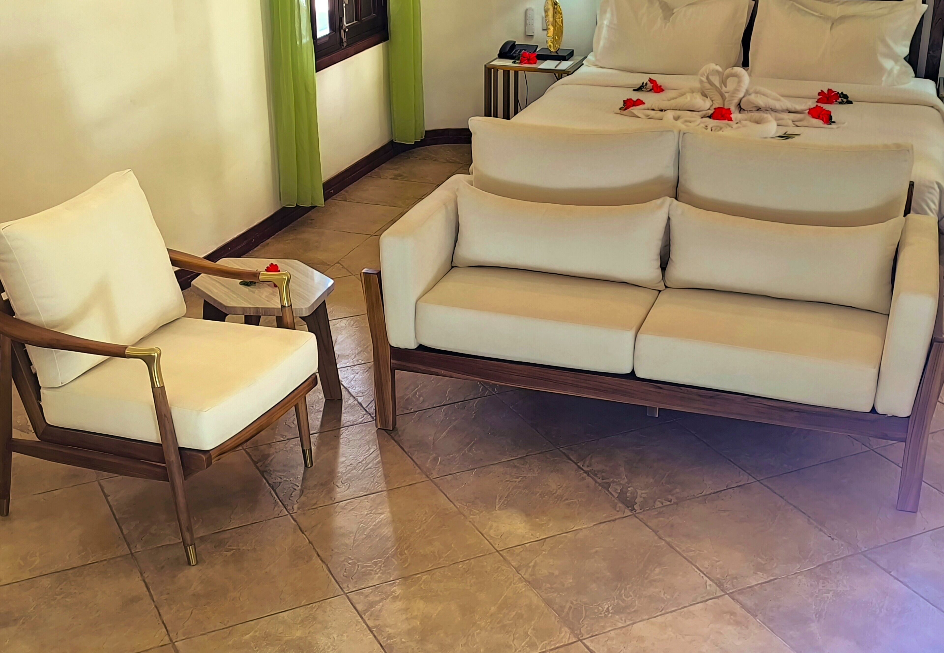 Habitación de hotel con un sofá blanco, un sillón de madera, una mesita y una cama con ropa de cama blanca y flores rojas.