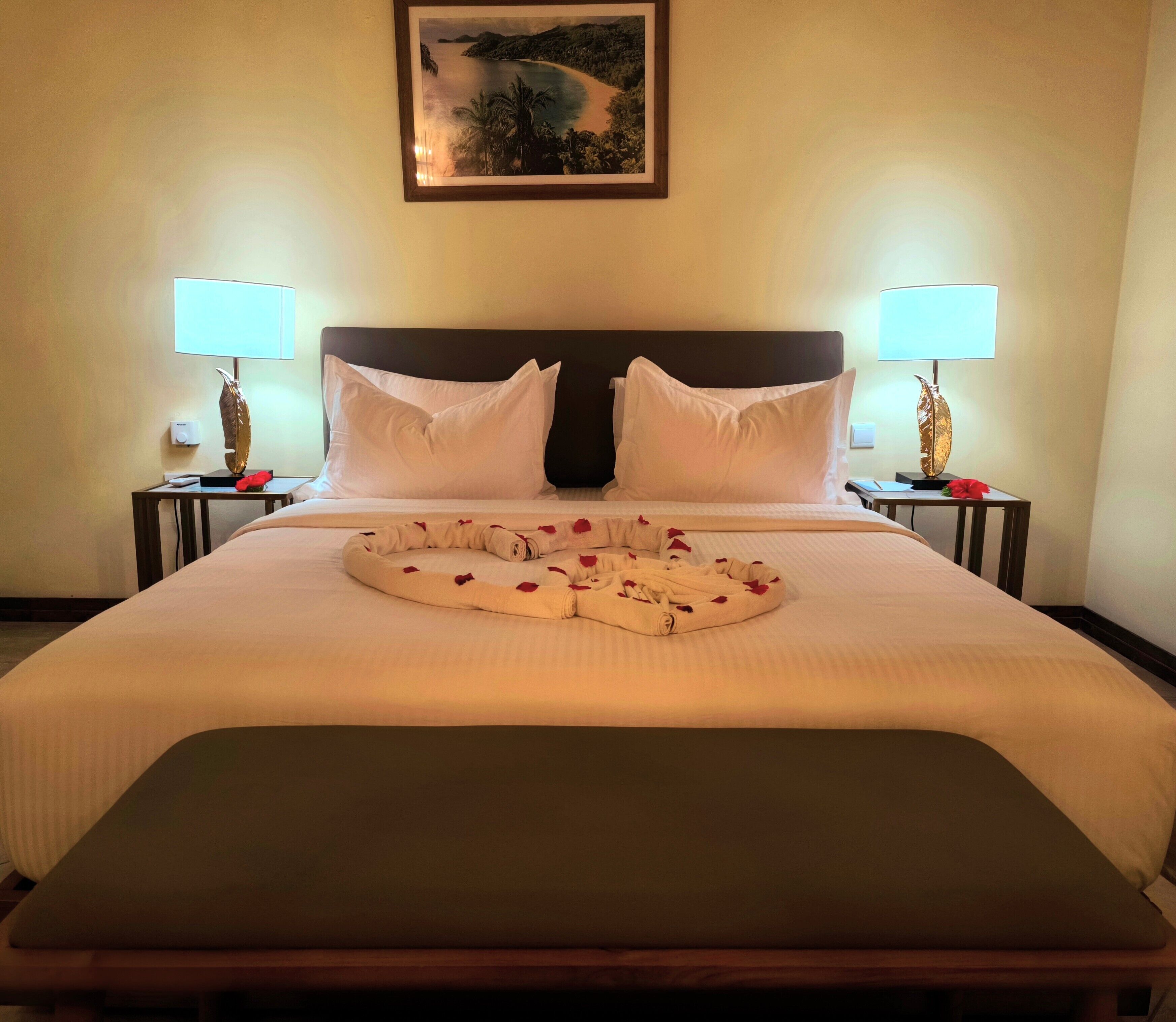 Een hotelkamer met een groot bed, twee nachtkastjes met lampen, een lijst aan de muur en een decoratieve kussenset op het bed.