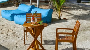 A wooden table with a bottle and a plate of food, a blue sofa, and a wooden chair on a sandy beach. - Castello Beach Hotel (Praslin Island)