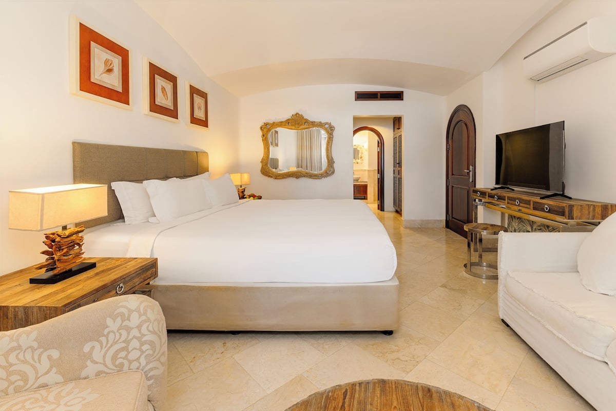 Quarto de hotel com uma cama grande, televisão, sofá, mesa de cabeceira de madeira e um espelho.