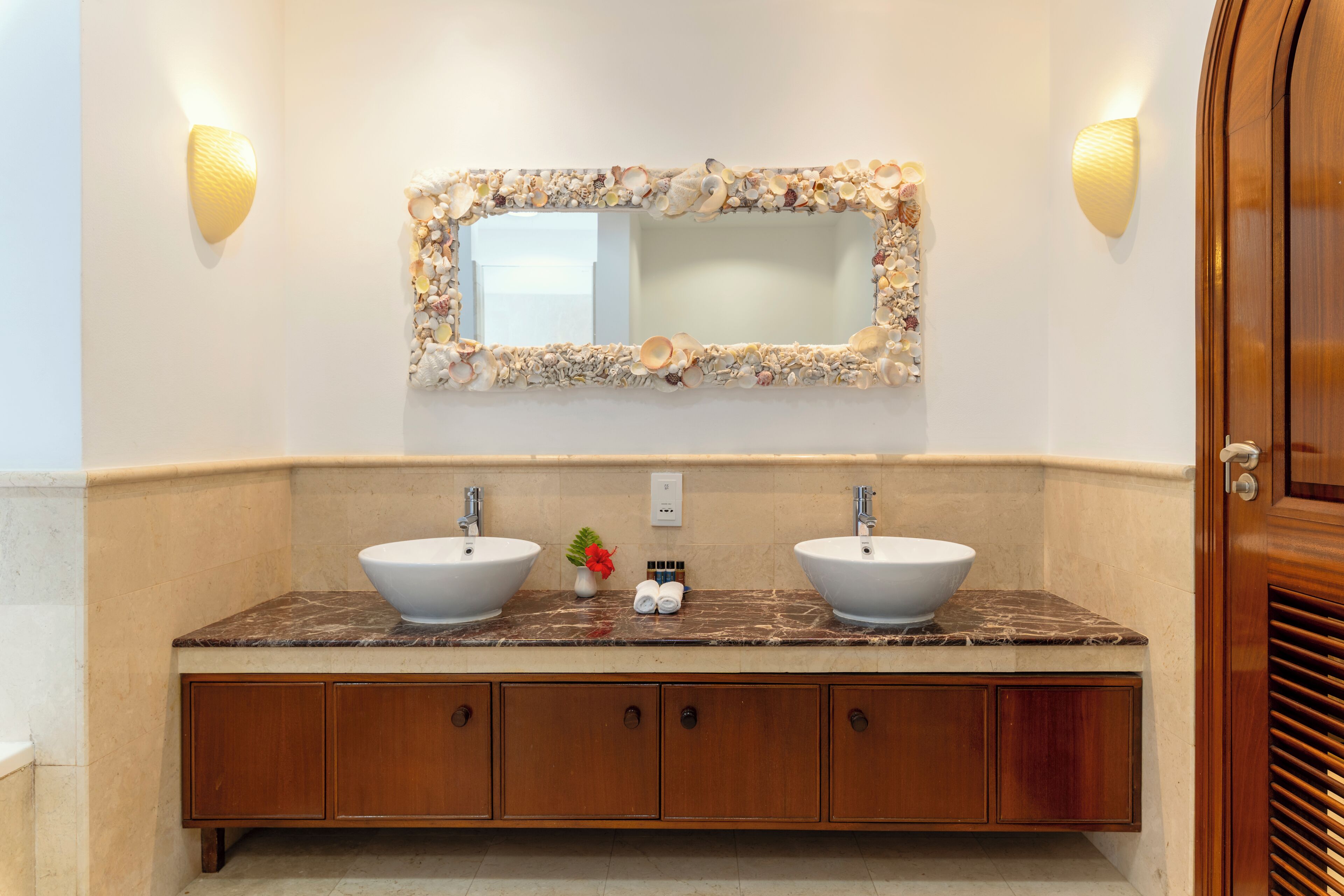 A bathroom with two sinks, a mirror with a shell frame, wooden cabinets, and wall sconces.