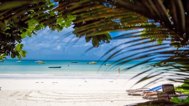 A beach with boats, a person walking, and a lounge chair. - Village Du Pecheur (Praslin Island)