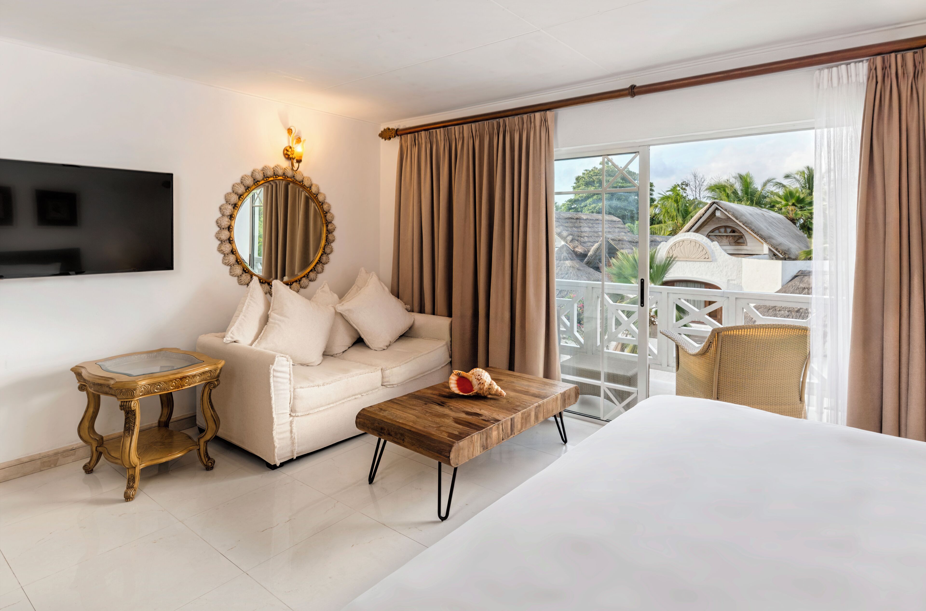 Habitación de hotel con cama, sofá, mesa de centro, televisor, espejo y balcón con mobiliario exterior.