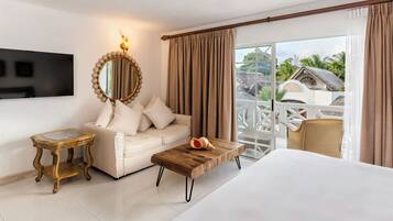 Habitación de hotel con cama, sofá, mesa de centro, televisor, espejo y balcón con mobiliario exterior.
