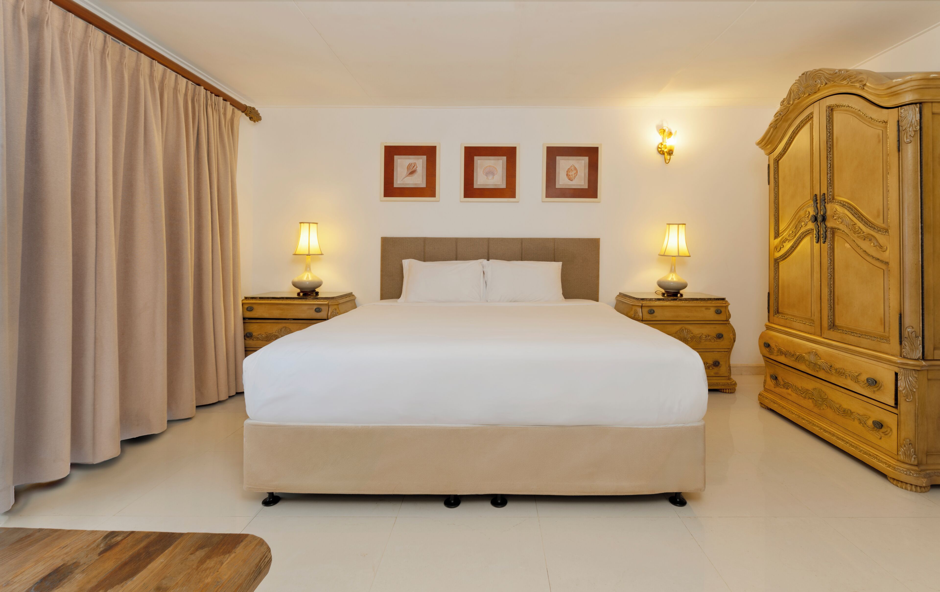 A hotel room with a large bed, two bedside tables with lamps, a wardrobe, and three framed pictures on the wall.