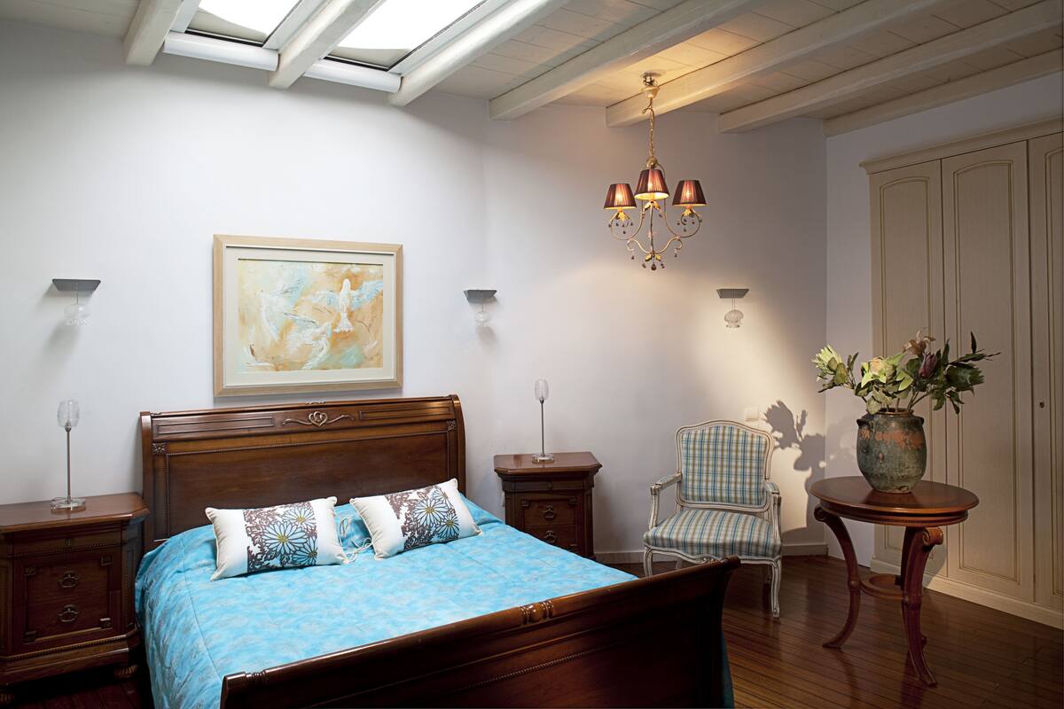 Une chambre à coucher avec un lit en bois, une couvre-lit bleue, un tableau au mur, un lustre, une chaise et une table sur laquelle se trouve un vase.