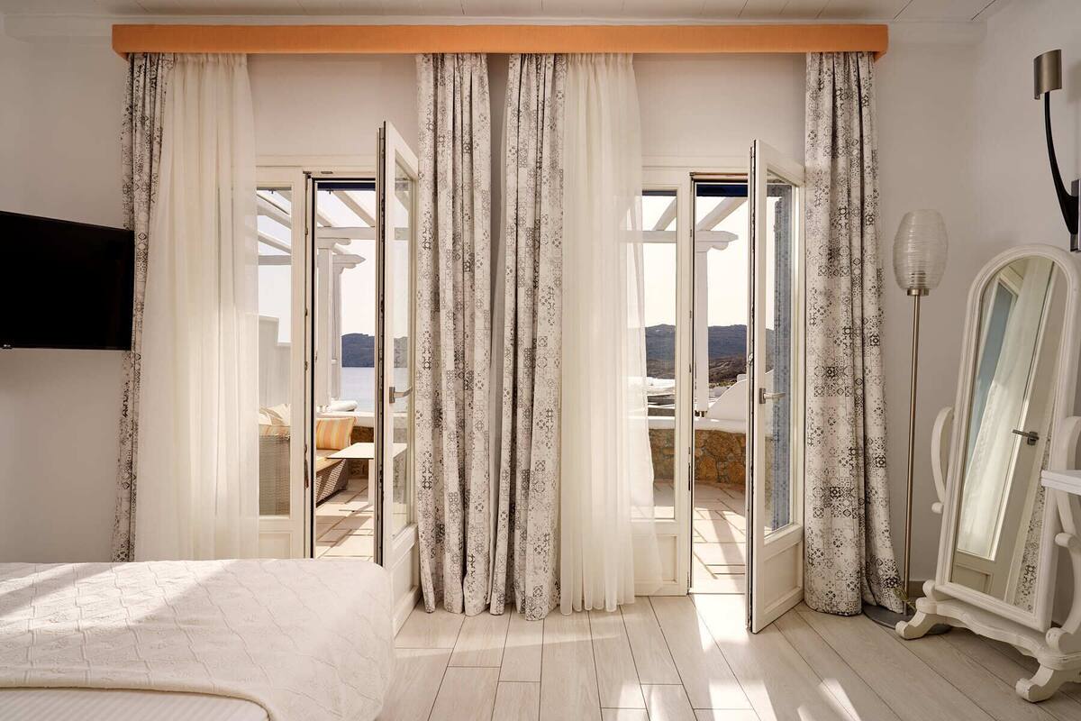 Une chambre à coucher avec un lit, une commode et un grand miroir. Les fenêtres sont dotées de rideaux, et on peut voir le paysage extérieur.