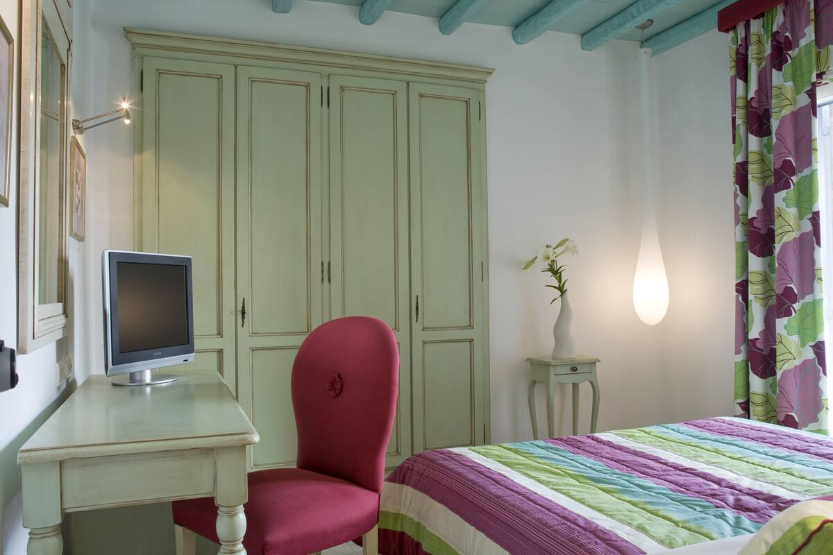 Une chambre à coucher avec un lit, un bureau avec un ordinateur, une chaise rouge, une armoire et une petite table sur laquelle est posé un vase.