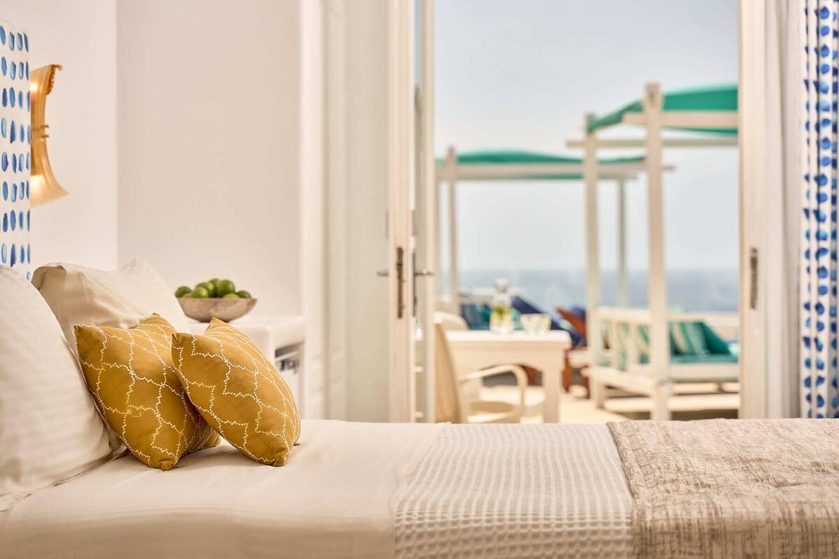 Une chambre à coucher avec un lit, deux oreillers, une petite table sur laquelle se trouve un saladier rempli de fruits, et une vue sur une terrasse avec une table et des chaises.