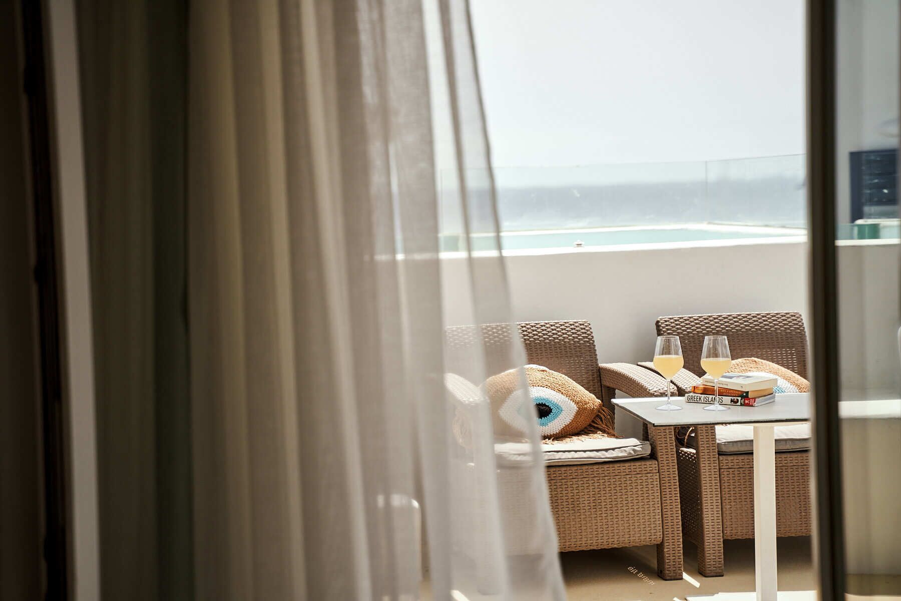 A balcony with wicker furniture, two glasses of white wine, and a stack of books titled "Greek Islands".