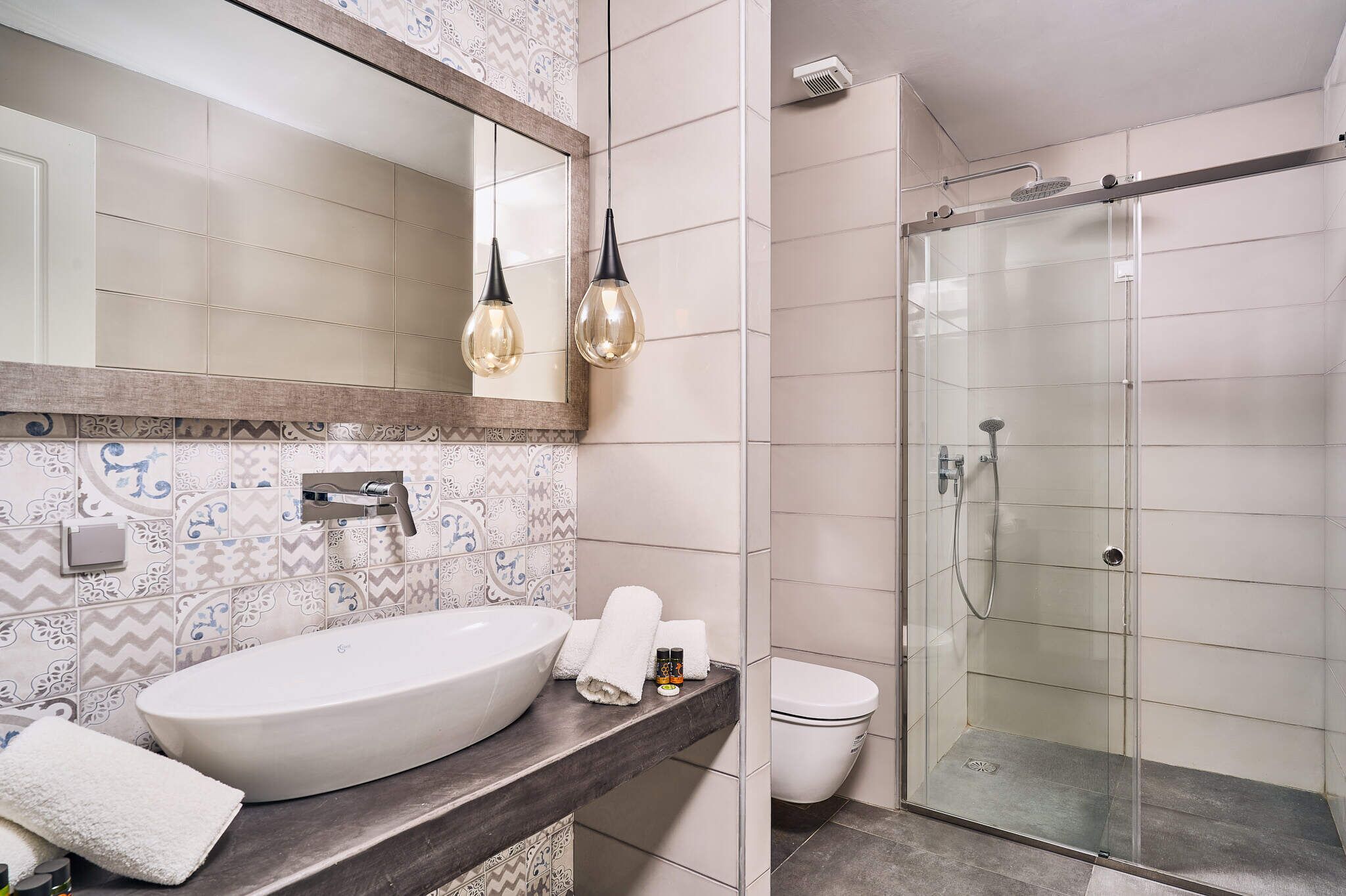 A bathroom with a large white sink, a mirror, two pendant lights, a shower, and a toilet.