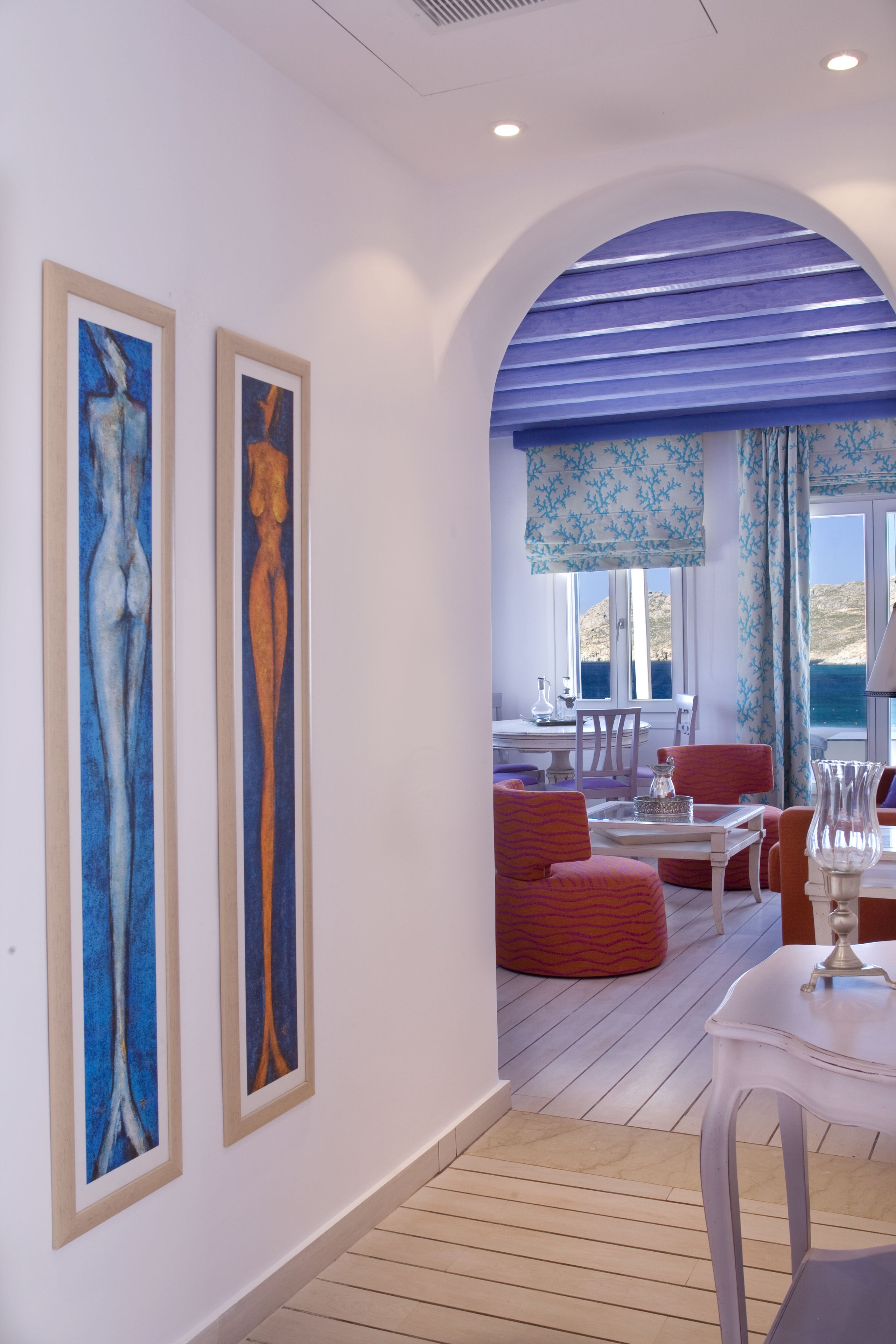 A modern interior with two framed artworks, a dining area with red upholstered chairs, and a view of the sea through large windows.