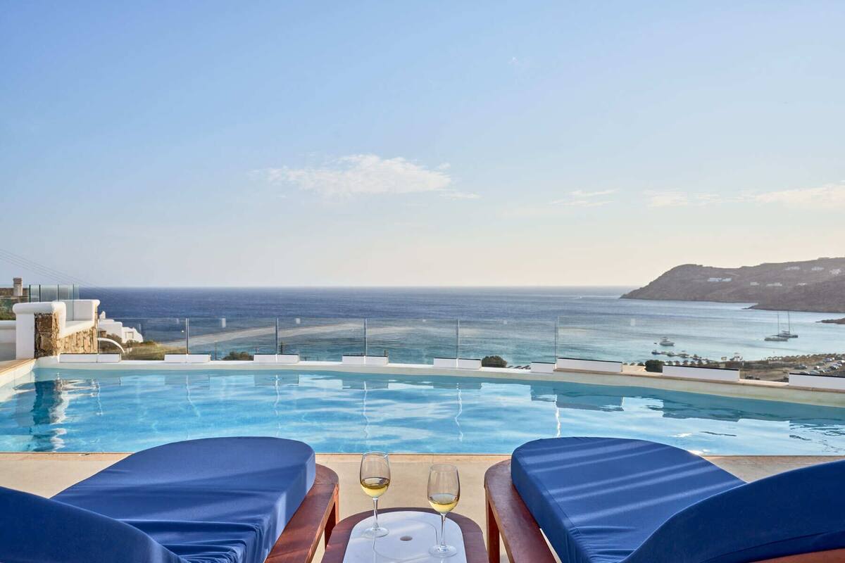 Une piscine à débordement avec deux chaises longues et une table, offrant une vue imprenable sur la côte, avec des bateaux et une île au loin.