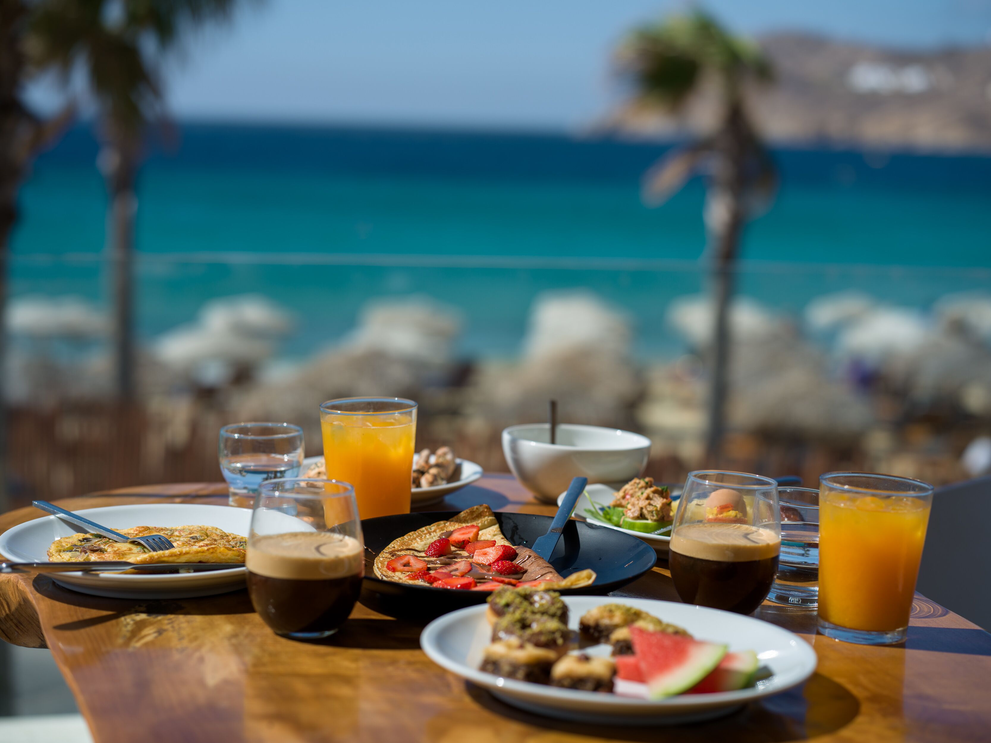 Una mesa con alimentos para el desayuno, como tostadas, fruta y bebidas, con vista a una playa de aguas azules cristalinas y palmeras.