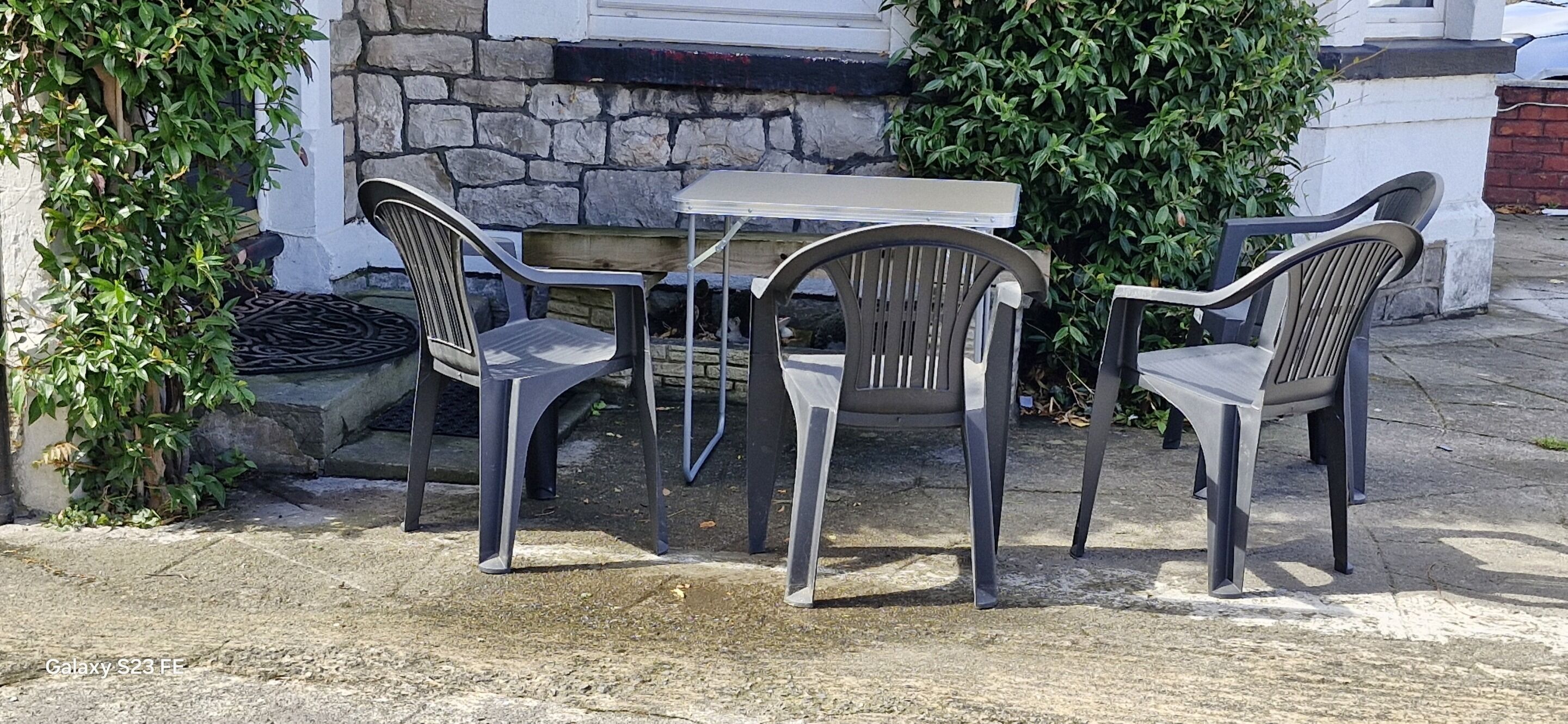 Un patio con tavolo e tre sedie, circondato dal verde e da un muro in pietra.
