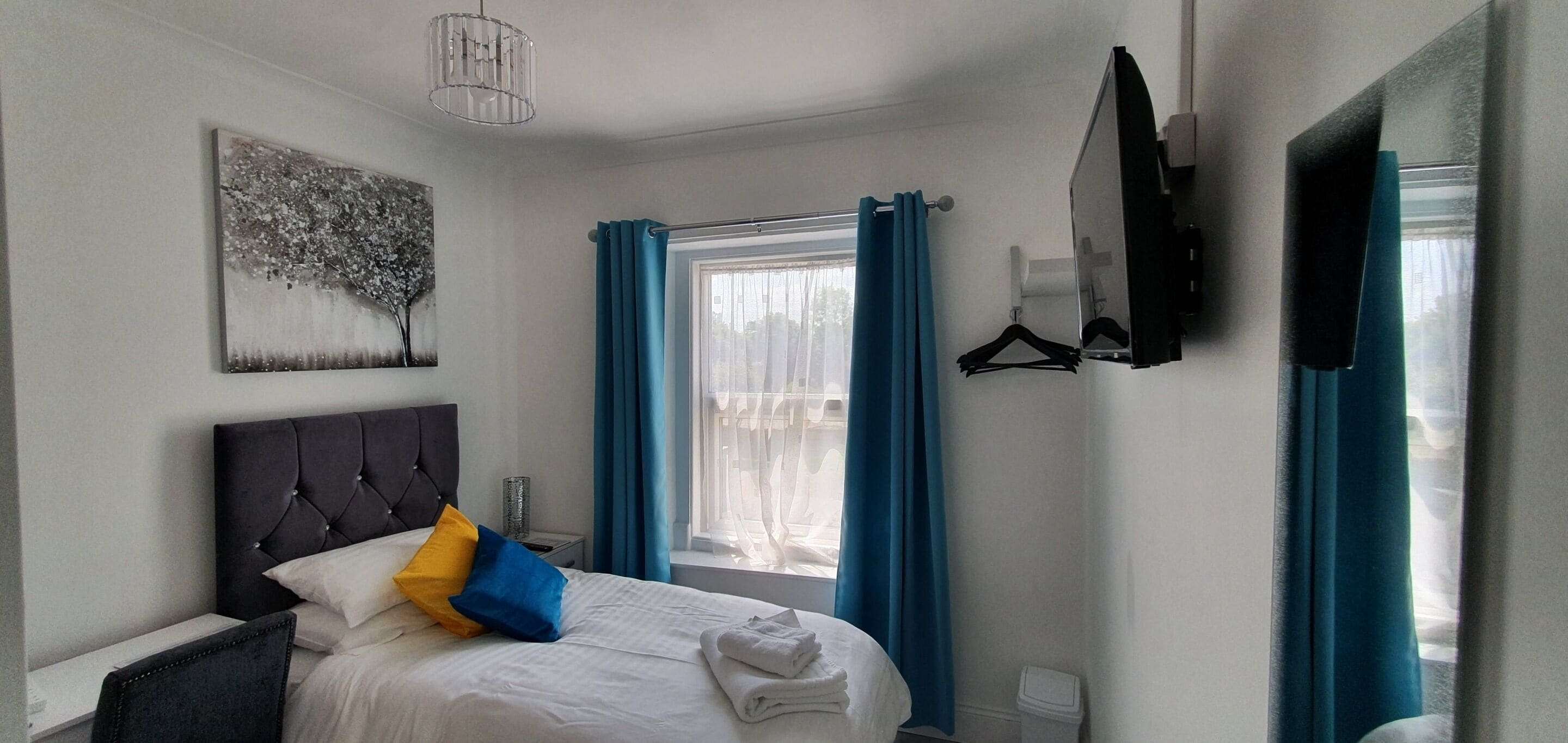 Une chambre d’hôtel avec un lit, une fenêtre avec des rideaux, un téléviseur fixé au mur et un tableau.