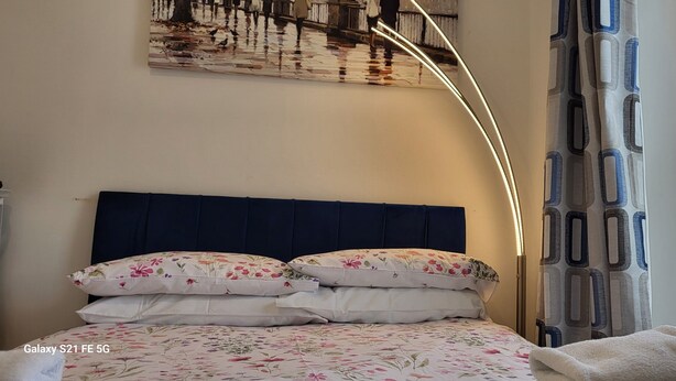 Een netjes opgemaakt bed met bloemenbedrukking, een donker hoofdbord en een wandlamp.