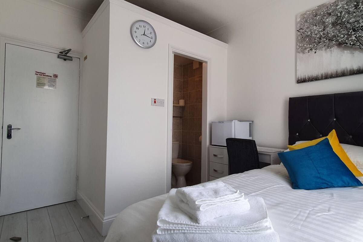 Habitación de hotel con cama, un reloj, un cuadro y un baño visible a través de una puerta abierta.