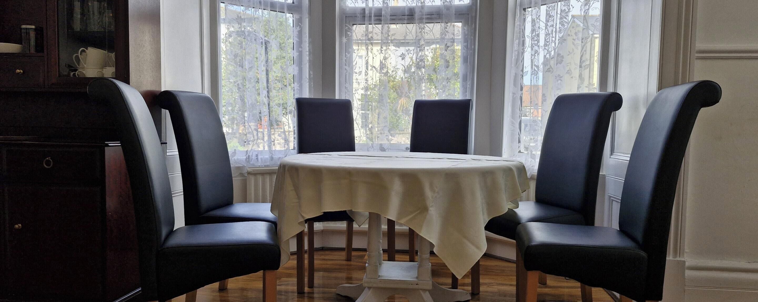 A dining area with a round table, four chairs, and a view through a window with sheer curtains.