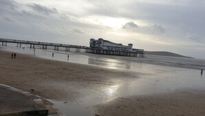 Een strand met een steiger die het water in loopt, een gebouw met meerdere verdiepingen, en een heuvelachtig landschap op de achtergrond.