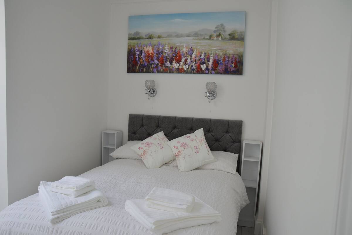 Una cama bien tendida con sábanas blancas, dos almohadones decorativos, un cabecero capitonado y un cuadro de un campo de flores sobre ella.