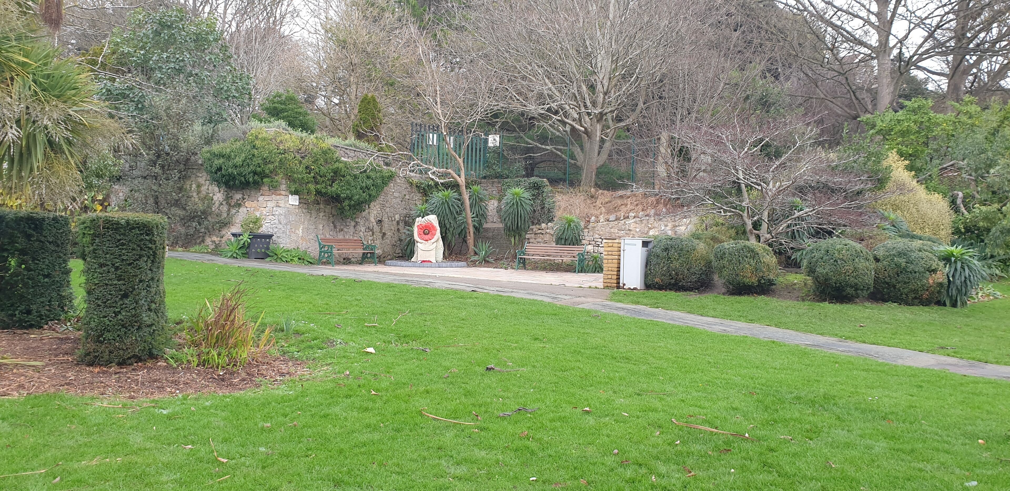 A park with a paved walkway, benches, and various shrubs and trees.
