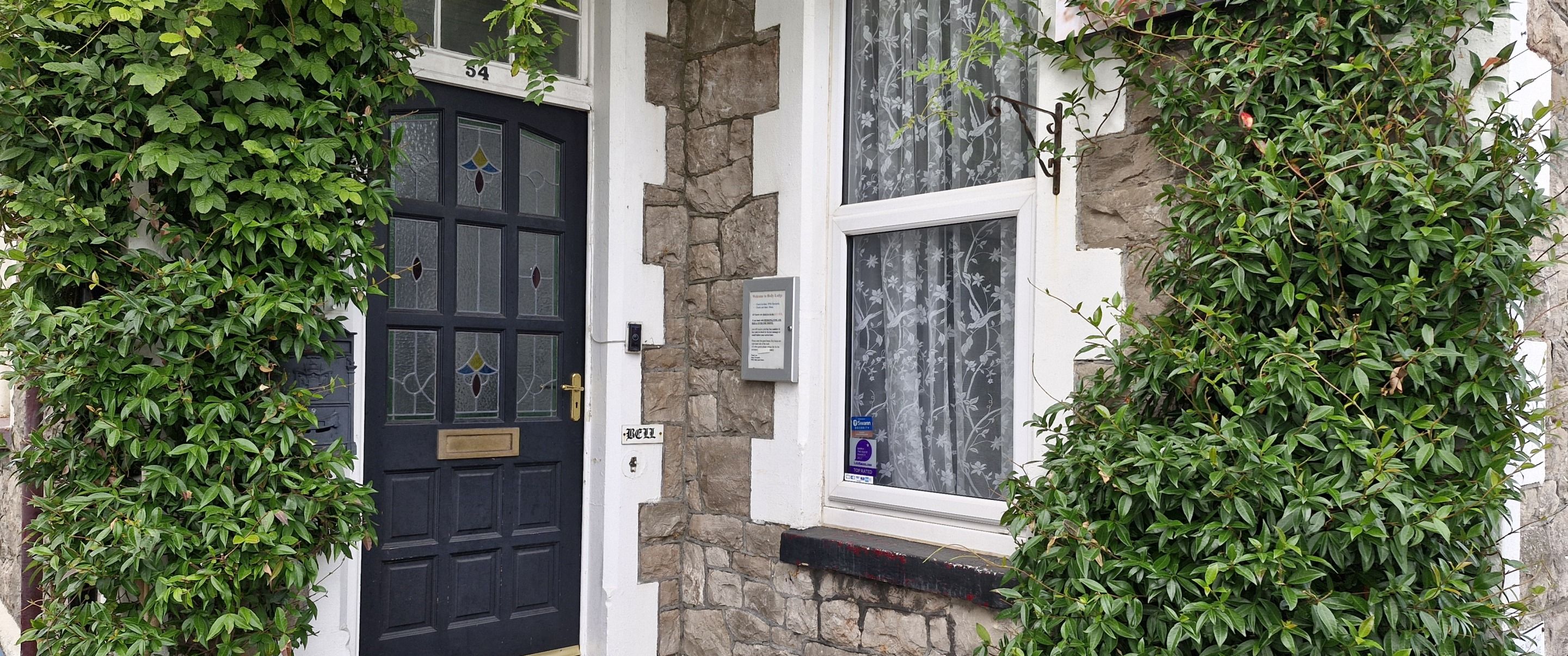A black door with a brass handle, a stone wall, and a window with floral curtains.