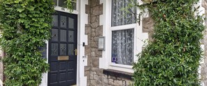 Eine schwarze Tür mit Messingklinke, eine Steinwand und ein Fenster mit floralem Vorhang.