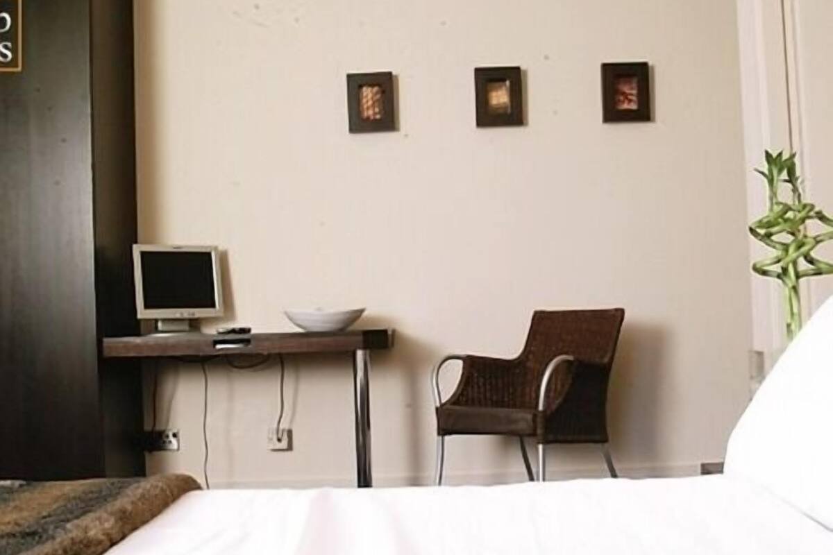 A hotel room with a bed, a desk with a computer monitor, a chair, and three framed pictures on the wall.