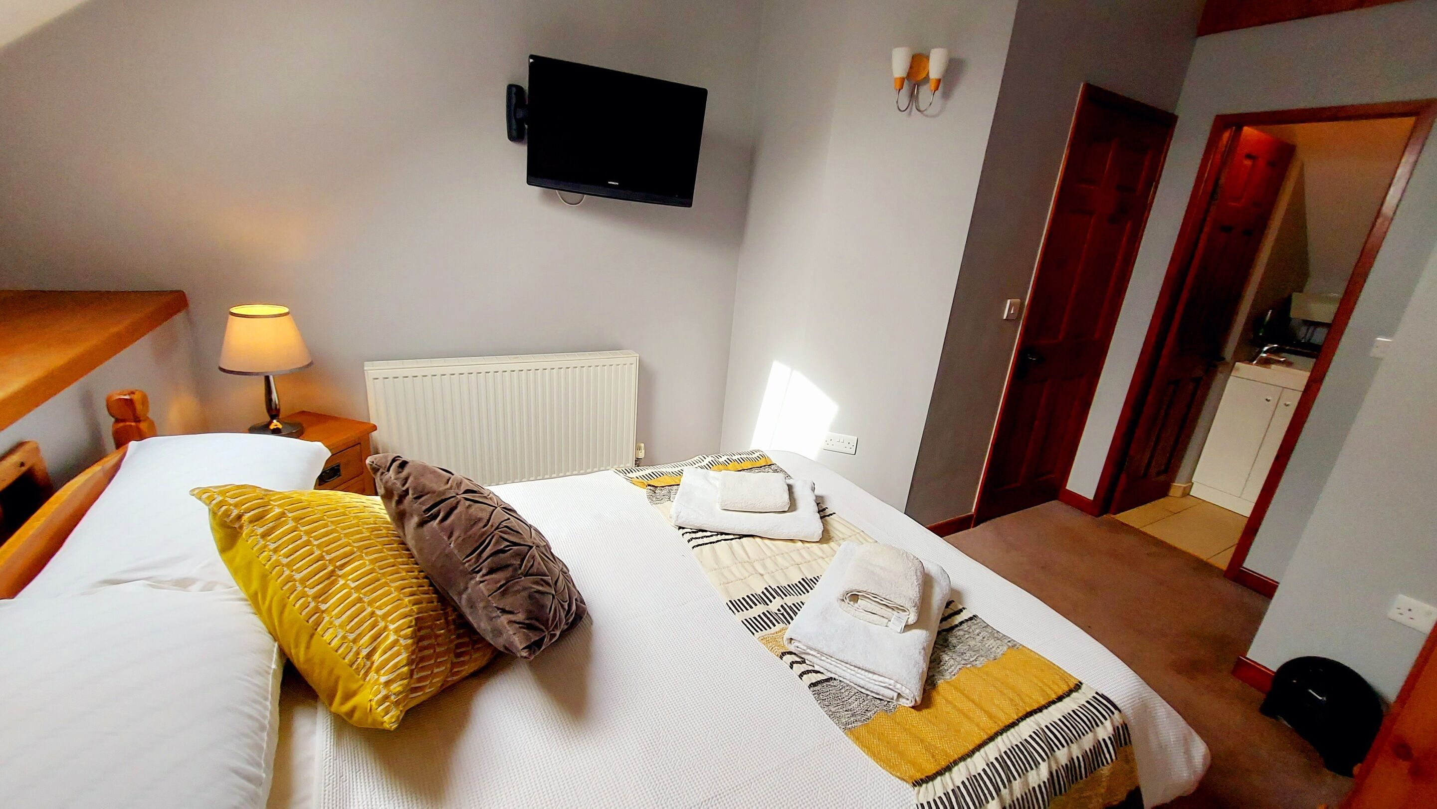 Een net opgemaakt bed met kussens, een bedlampje, een flatscreen-televisie aan de muur en een deur naar een badkamer.