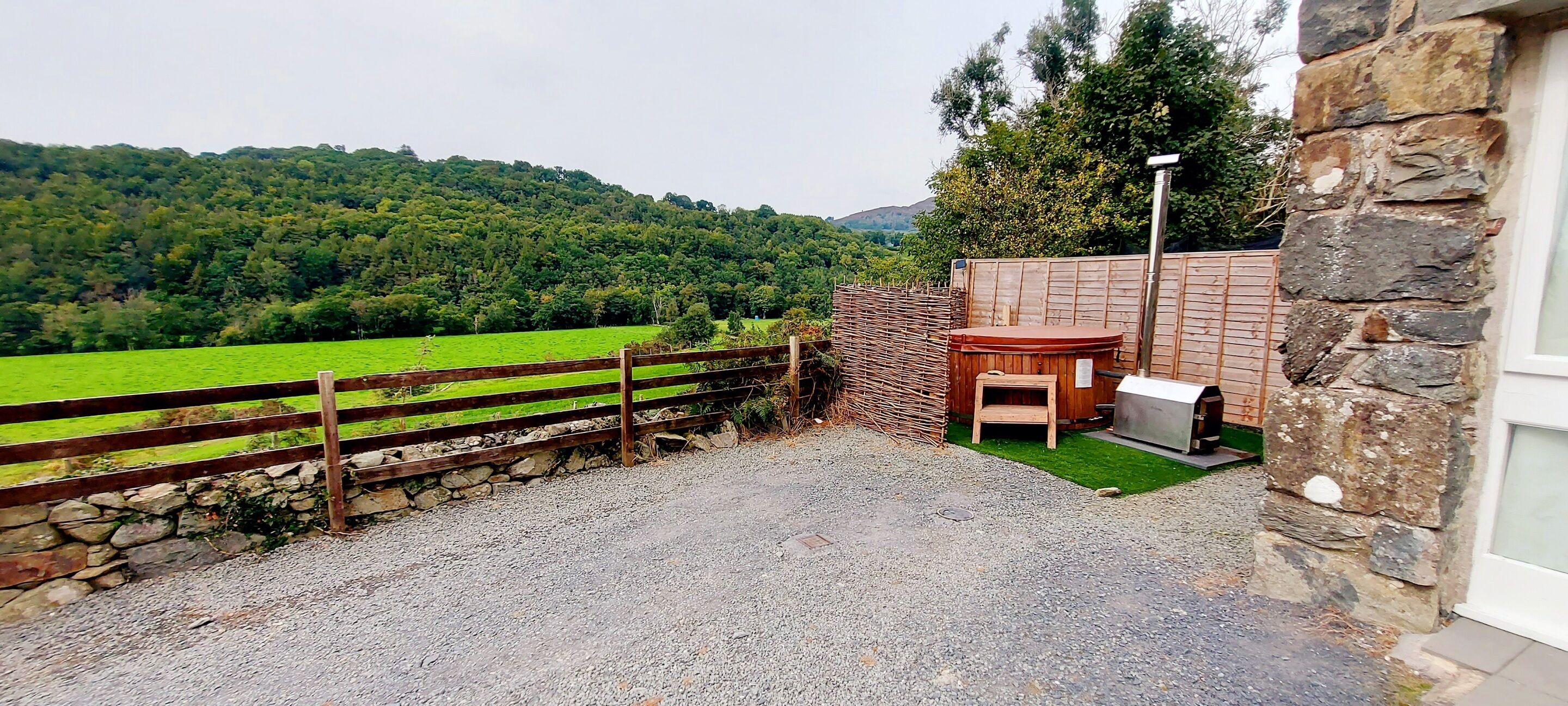 A patio area with a stone wall, a wooden fence, and a view of a green field and forested hills.