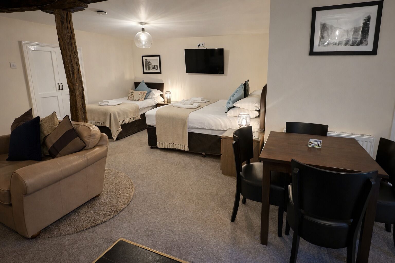 A compact hotel room with two single beds, a sofa, a dining table with chairs, a TV, and a framed picture on the wall.