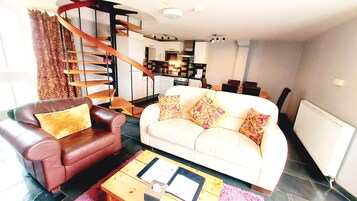 A modern living room with a brown leather sofa, a white sofa with floral cushions, and a wooden coffee table. A spiral staircase leads to an upper level with a kitchen area.