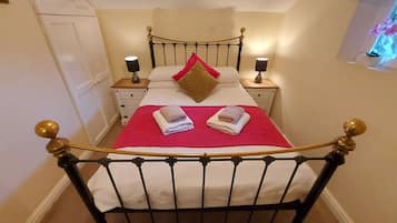 Una cama bien hecha con una colcha roja y dos toallas dobladas, una mesita de noche con una lámpara y una mesilla con un jarrón de flores.