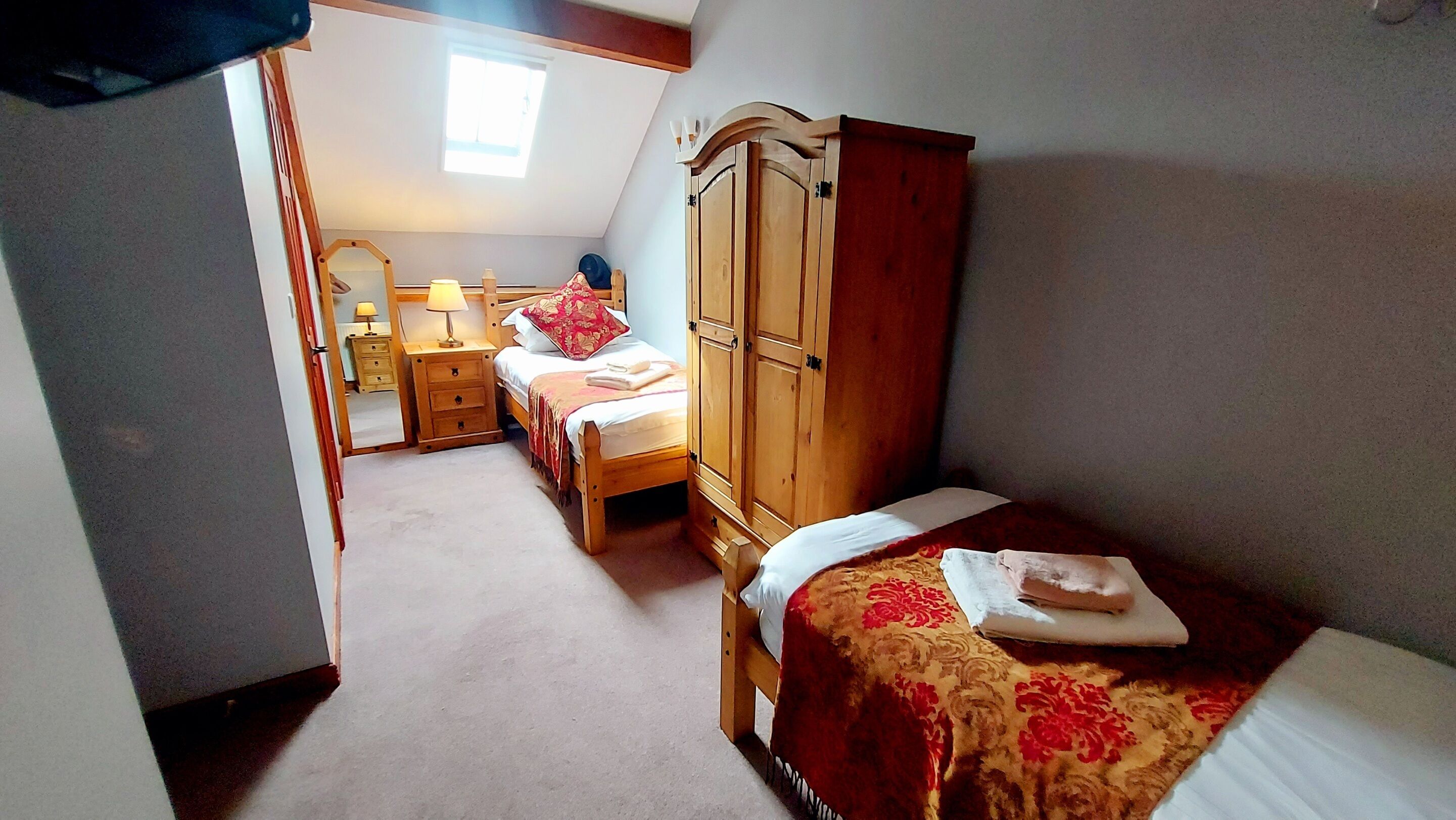A bedroom with two single beds, a wooden wardrobe, a bedside table with a lamp, and a mirror.