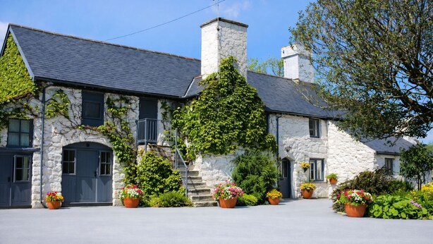 A stone building with a grey roof, a chimney, and a staircase leading to a balcony. There are potted plants in front of the building.