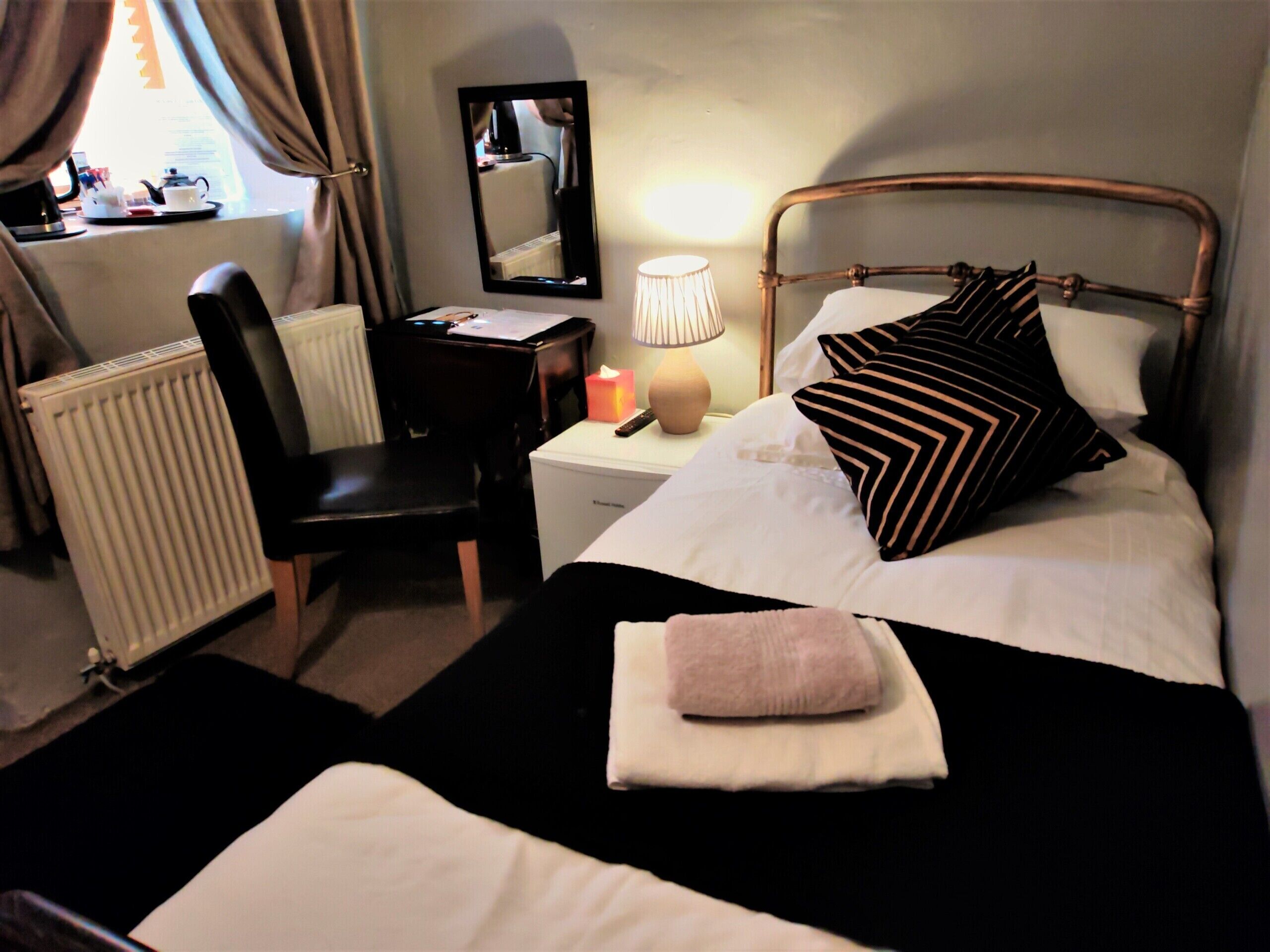 A hotel room with a bed, a desk, a chair, a radiator, a lamp, and a mirror.