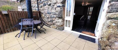 A stone building with a patio area featuring a table and chairs, a wooden fence, and a door leading inside.