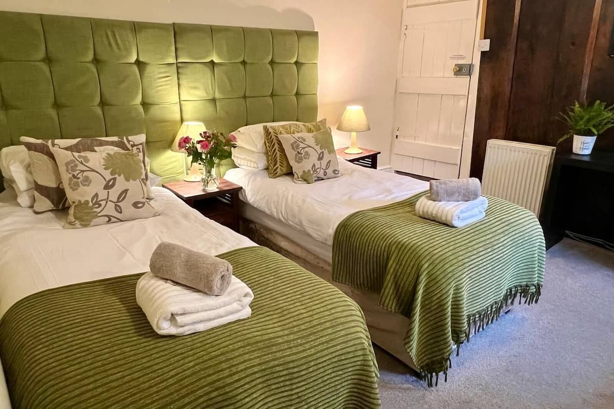 Una habitación de hotel con dos camas, cabecera verde, lámparas de noche y una planta en maceta.