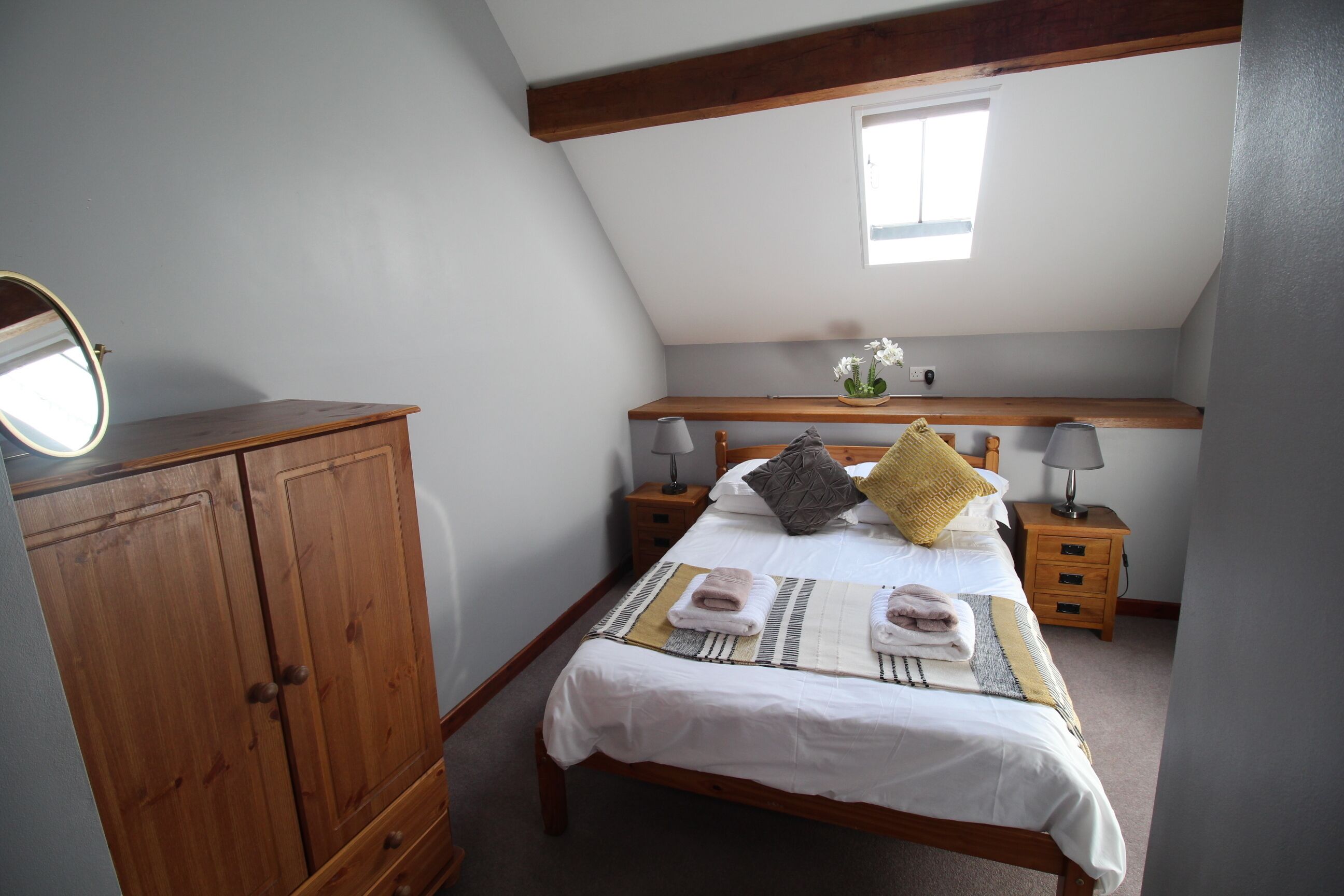 A bedroom with a wooden wardrobe, a bed with white bedding, two bedside tables with lamps, a window, and a small shelf with flowers.