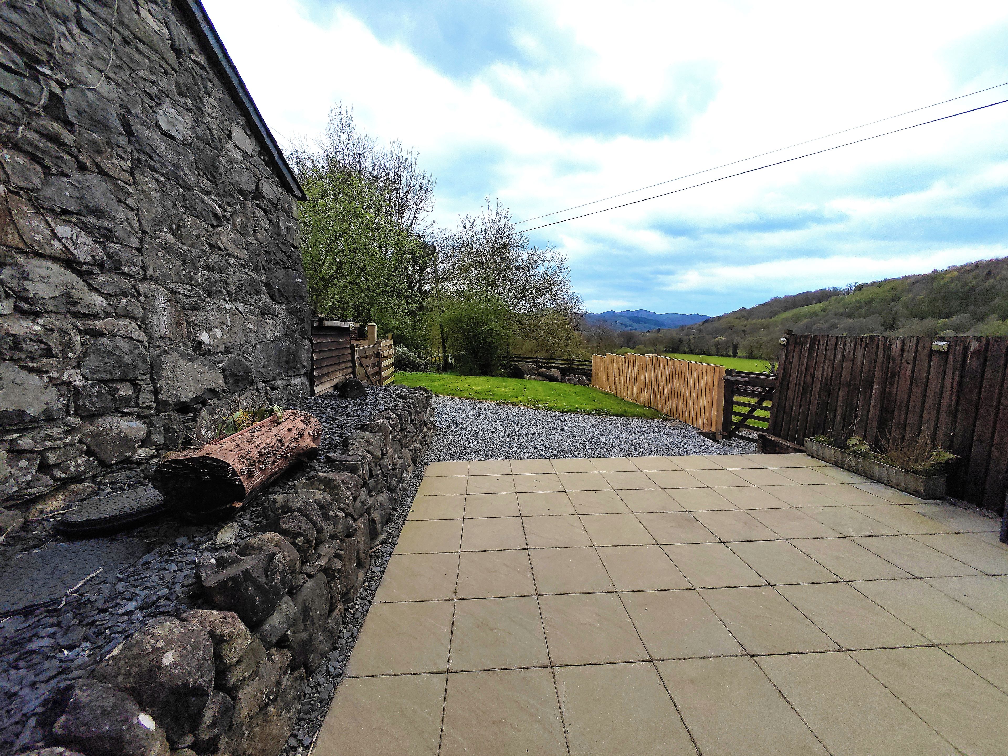 A stone wall, a wooden fence, and a paved patio leading to a grassy area with trees and hills in the background.