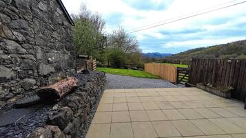 A stone wall, a wooden fence, and a paved patio leading to a grassy area with trees and hills in the background.