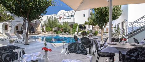 Comedor con mesas dispuestas para dos, piscina al aire libre y edificios blancos con arcos al fondo.