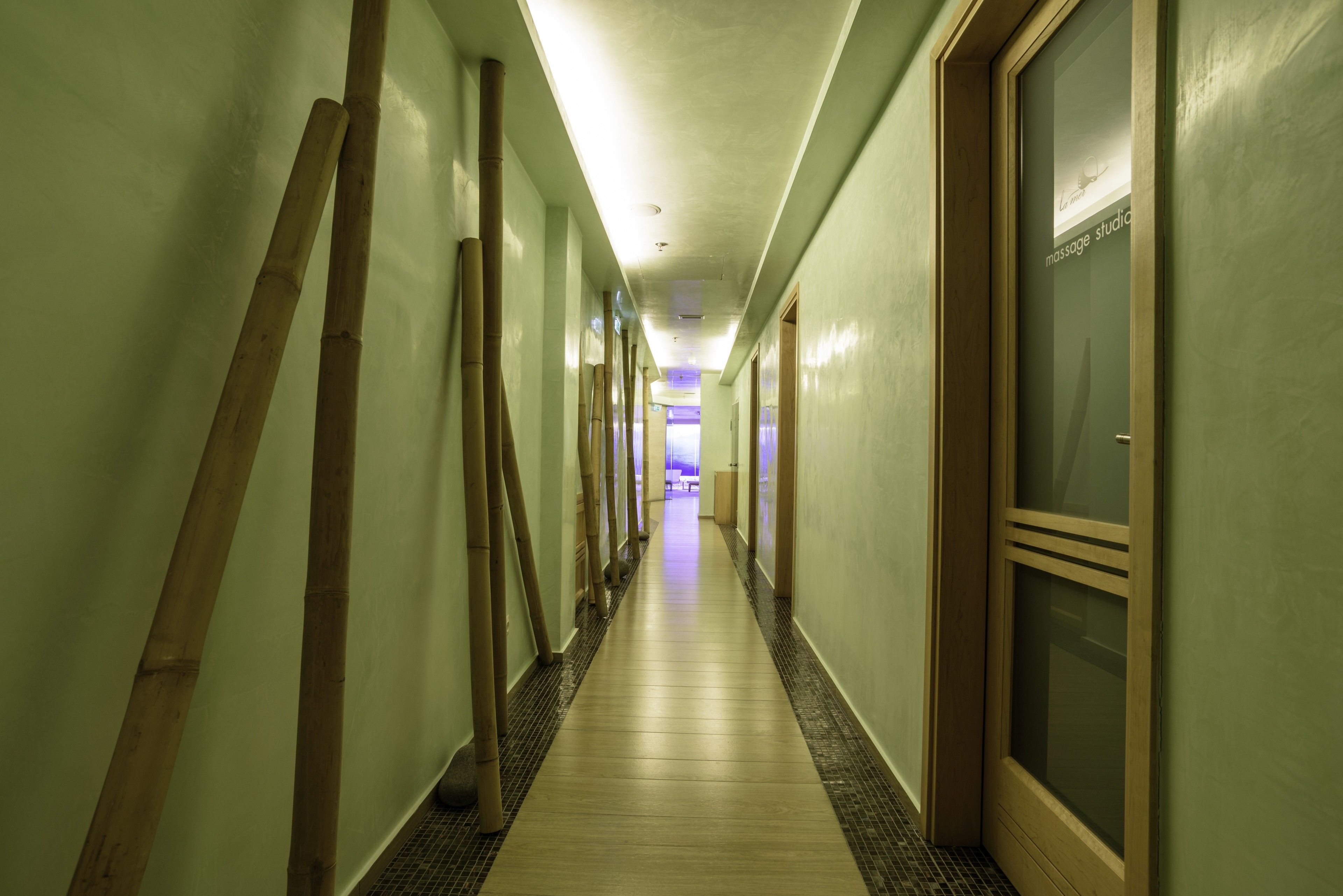 A hotel corridor with a wooden floor, green walls, and a massage studio door at the end.