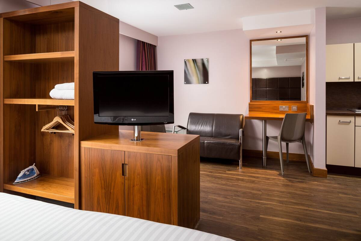 A hotel room with a wooden wardrobe, a flat-screen TV, a sofa, a dining table with chairs, and a kitchen area.