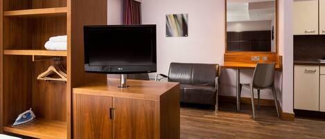 Hotellihuone, jossa on puinen vaatekaappi, taulu-tv, sohva, ruokapöytä tuoleineen ja pieni keittiö.