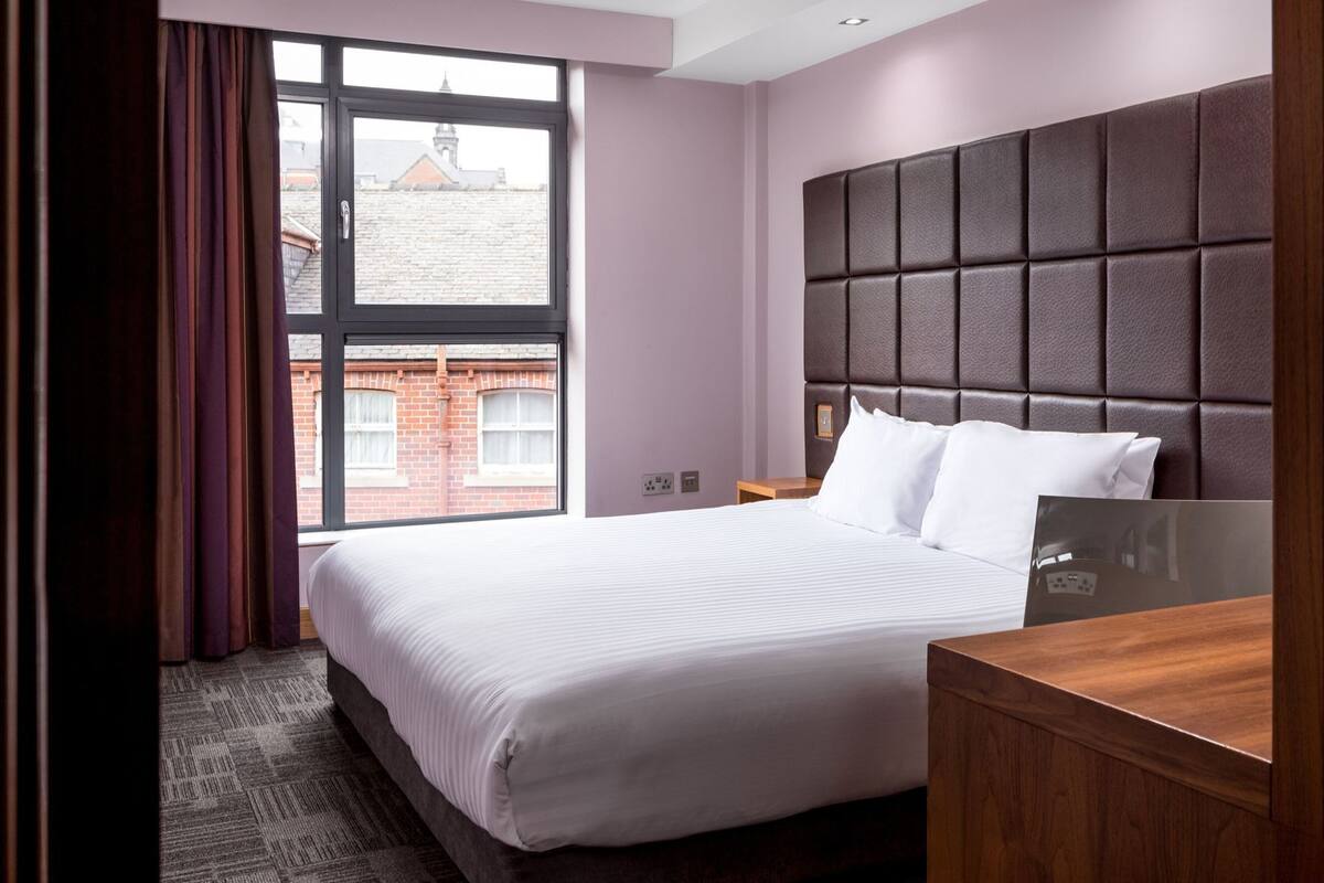 A hotel room with a large bed, a headboard, a wooden nightstand, a mirror, and a view of a brick building through a window.