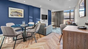 Una habitación de hotel moderna con una pared de acento azul, una zona de comedor con una mesa redonda y sillas, un sofá, un televisor montado en la pared y un ventanal con cortinas.