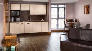 A modern kitchen with a microwave, toaster, and coffee maker. There is a brown leather sofa and a glass table with a laptop.